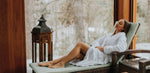 A woman in a white bathrobe relaxes on a wicker lounge chair by a large window overlooking a wintery forest.