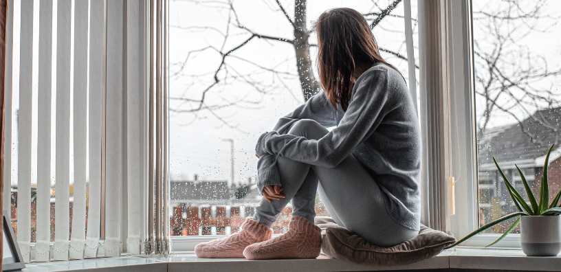 A woman sits by a window, gazing thoughtfully outside as rain drizzles on the glass, creating a cozy yet sad atmosphere.