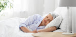 An older woman with gray hair sleeping contently in bed, in a light-filled room, symbolizing serene sleep and the importance of restorative rest for wellness in later years.