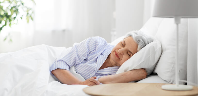 An older woman with gray hair sleeping contently in bed, in a light-filled room, symbolizing serene sleep and the importance of restorative rest for wellness in later years.