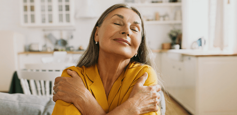Older woman in yellow blouse hugs herself in her home