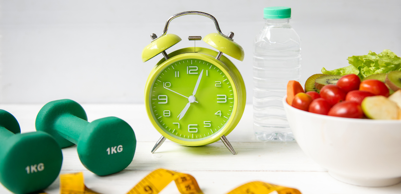 Still life image of a green alarm clock, dumbbells, water bottle, fruit bowl, and measuring tape on a white surface.