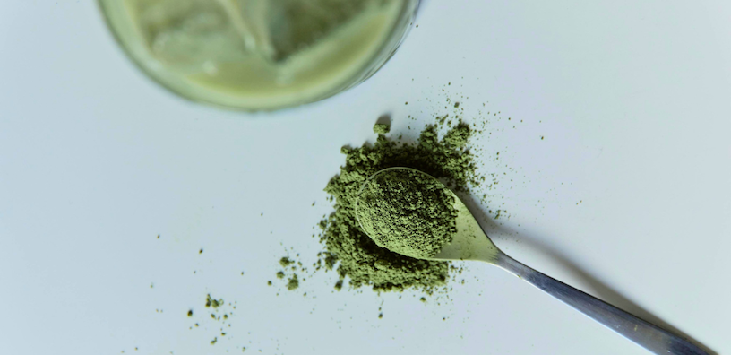 A spoon overflowing with vibrant green powder on a white surface. A glass with a green drink is partially visible in the top corner.