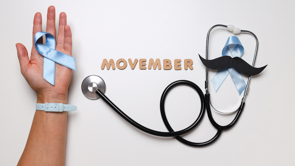 A hand holds a blue awareness ribbon while a stethoscope and paper mustache lie beside wooden letters spelling “Movember.”