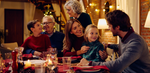 A multi-generational family enjoys a festive holiday meal around a decorated dining table. Smiling parents, grandparents, and children sit together, with candles, pinecones, and string lights creating a cozy, celebratory atmosphere.