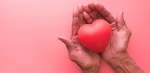 Elderly hands cupping a smooth, red, heart-shaped object against a pink background.