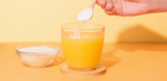  Hand scooping collagen powder into a glass of orange juice on a yellow background.