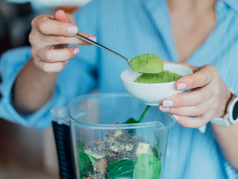 A woman carefully adding green powder into a blender, indicating a focus on health and nutrition.