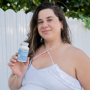 Woman holding a bottle of supplements outdoors with a white fence and greenery in the background