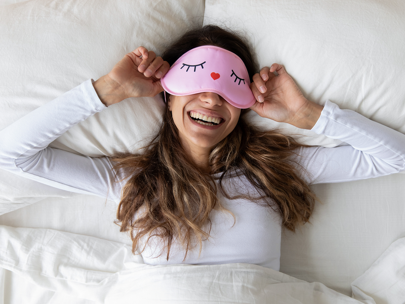 A woman smiles brightly while wearing a soft pink sleep mask.