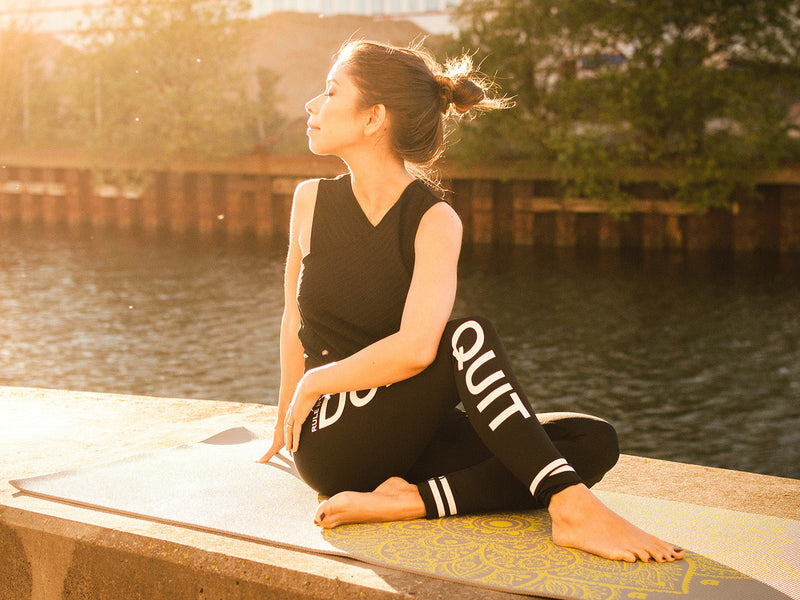 A woman sits on a yoga mat, peacefully practicing in front of a serene body of water.