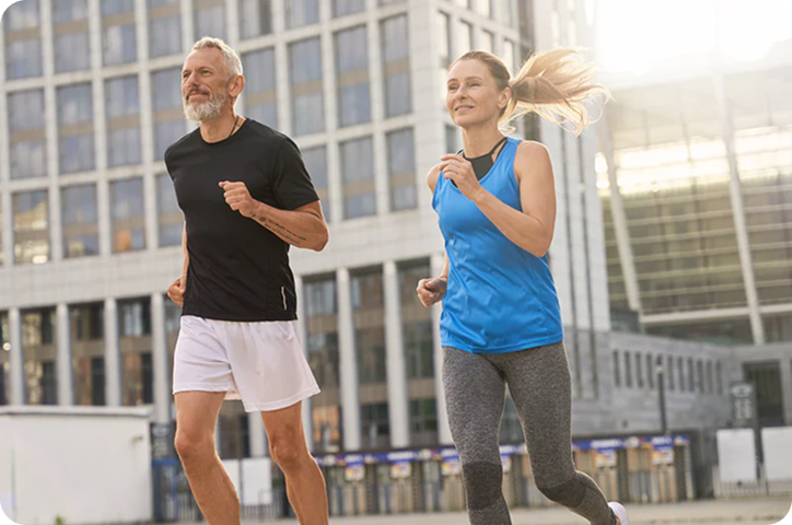 A mature man in a black shirt and a woman in a blue tank top jogging side-by-side with city buildings in the background.